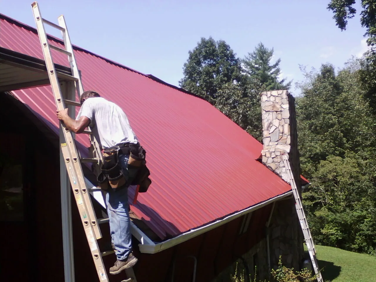 Roofer on a ladder beside a red metal roof and stone chimney — Soffit & Fascia Repair by Roofing Brothers in Clarkrange